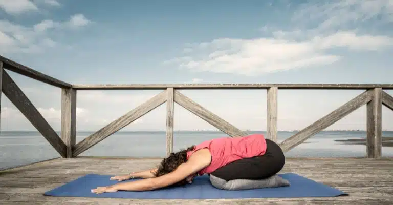 A woman in a red shirt and black pants practicing yoga in Child's Pose (Balasana), at a beach deck.