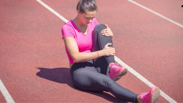 A female sports athlete experiences an injury during her game. She lies on the ground in pain, clutching her leg, while her teammates and opponents gather around her.