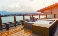 A serene view from a wooden deck featuring a hot tub, overlooking a calm lake with the majestic Mount Fuji in the background under a cloudy sky.