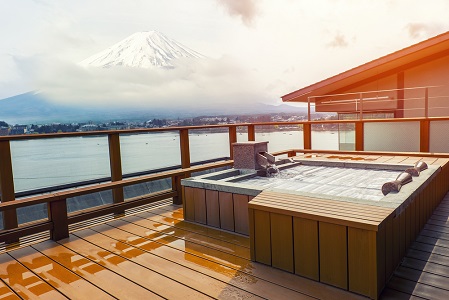 A serene view from a wooden deck featuring a hot tub, overlooking a calm lake with the majestic Mount Fuji in the background under a cloudy sky.