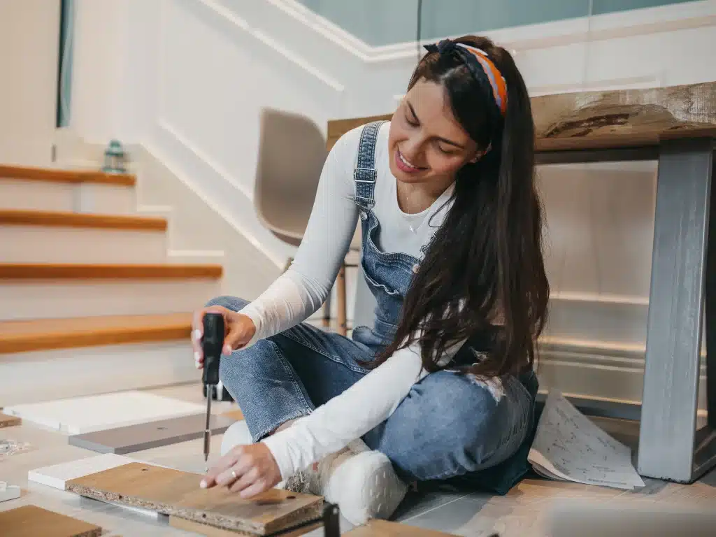 A woman engages in a DIY project, skillfully using a drill on a wooden board near a staircase, illustrating the hands-on approach to creating a custom hot tub table.