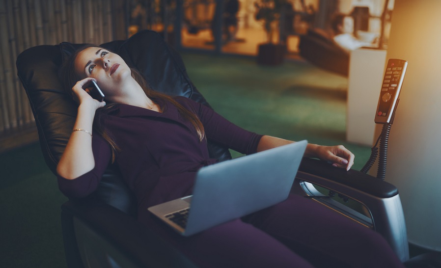 A woman sitting on a massage chair with a laptop on her lap and a phone in her hand.