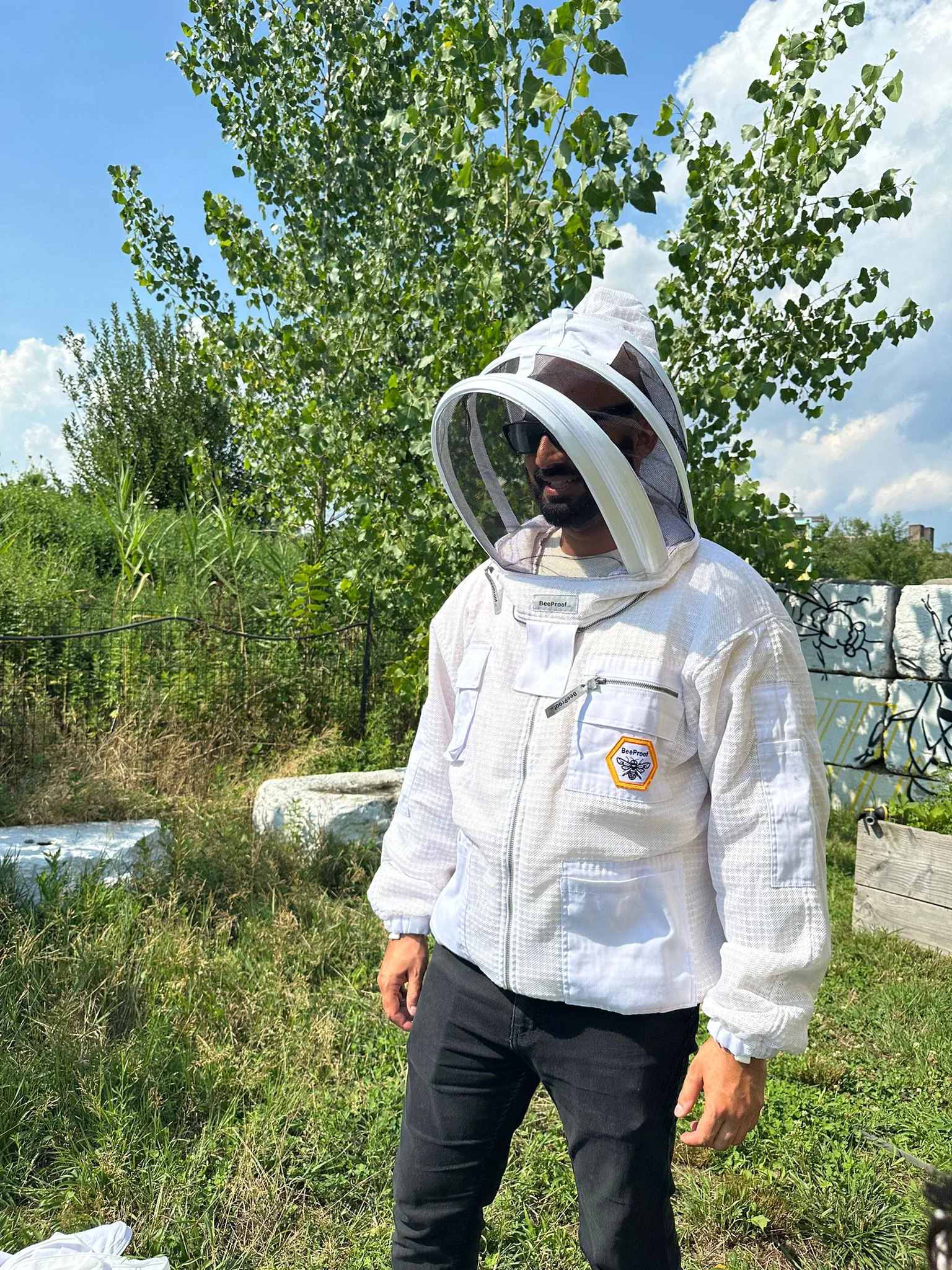 A person wearing a beekeeping suit with a veiled hat stands outdoors. The suit is white, and the individual is also wearing black pants. In the background, there are green trees and shrubs under a blue sky with scattered clouds.