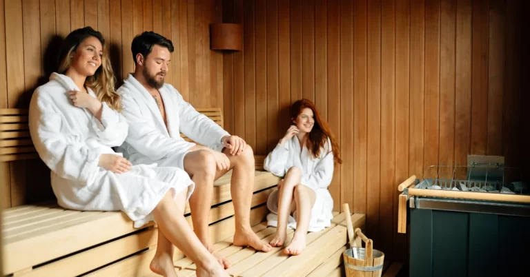 Three friends sitting inside a wooden sauna room. The individuals are wearing white robes and have their faces obscured by brown pixelation for privacy.