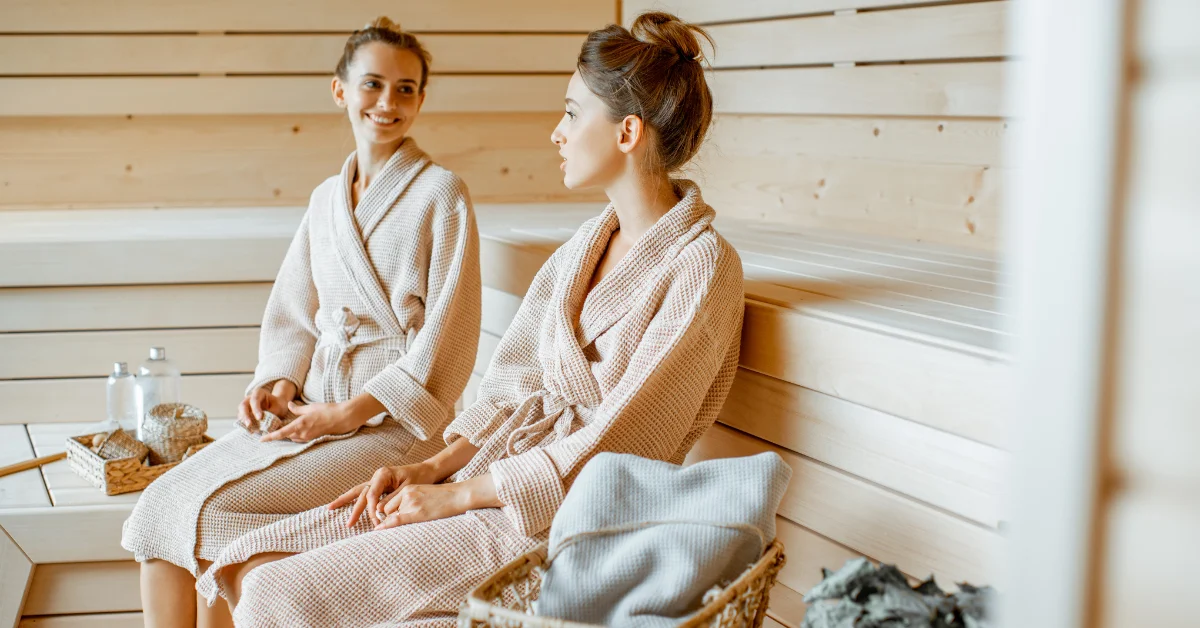 Two individuals sitting side by side on a wooden bench inside a sauna room, wearing light-colored bathrobes and with towels draped over their legs. Their faces are obscured for privacy. A basket of stones and a water bottle are visible on the floor beside them.