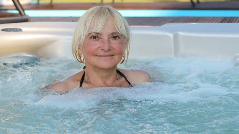 A senior citizen is relaxing in a white-hot tub with water jets and bubbles, surrounded by a pool and a blue sky. (Hot tubs for senior citizens)