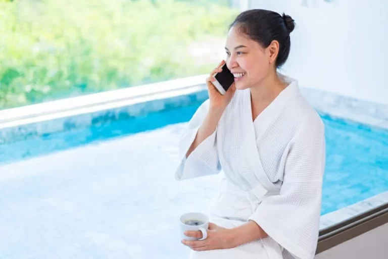 A young woman wearing a white robe holds a cup of coffee, preparing to use the hot tub in the morning.