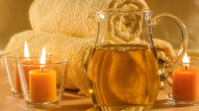 A glass bottle of massage oil, a wooden spoon, a bowl of dried flowers and a towel on a wooden table