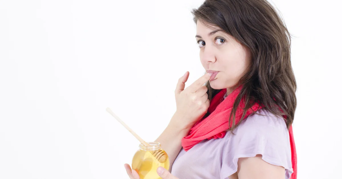 A person holding a jar of honey with a wooden dipper in it. The person’s face is blurred and they are wearing a pink shirt and a red scarf. The background is white.