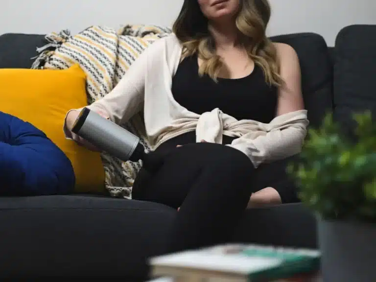 Well-dressed young woman enjoying a massage from a gun massager while seated on a sofa.