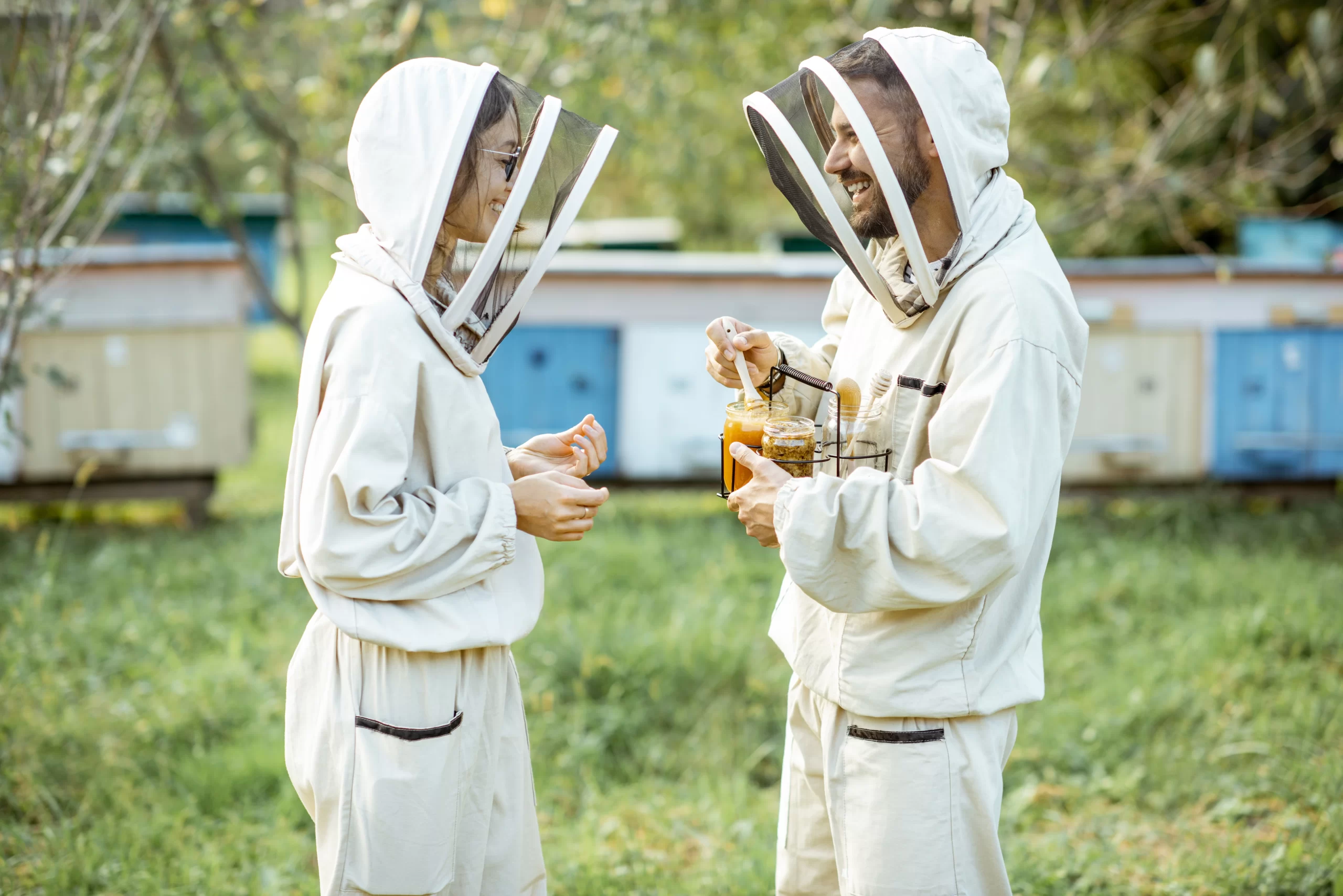 Two beekeeper in protective beekeeping suits are standing in front of several beehives. One individual is holding a frame with honeycombs, while the other is gesturing towards it. The environment suggests a lush green area, possibly a garden or farm dedicated to beekeeping.