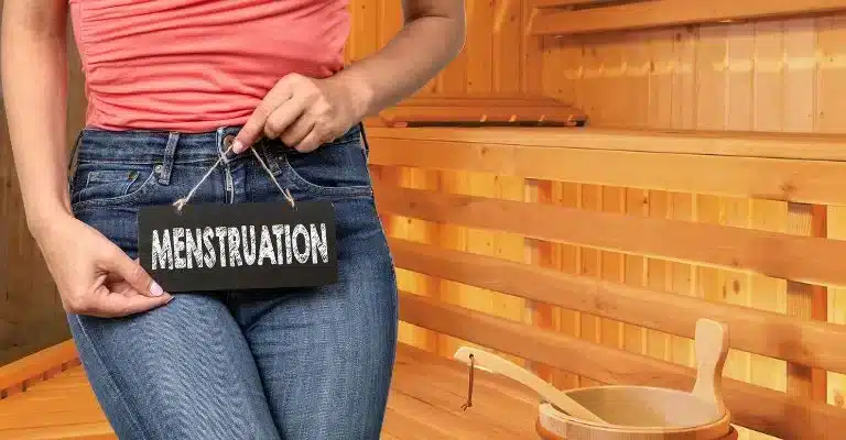 A woman holding a pouch with the word “MENSTRUATION” on it in a sauna. (Sauna During Menstruation)