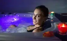 A woman in a blue-lit soaking in a hot tub with candles and bubbles at night.