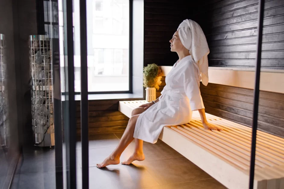 A person sitting on a wooden bench in a sauna, wearing a white robe and with a white towel on their head, with dark wooden walls and a window on the left side, and a metal rack with towels. (How Much Weight Can You Lose in a Sauna)