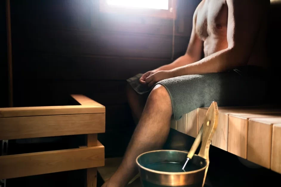 A person sitting in a sauna on a wooden bench with a towel draped over their lap, wearing gray shorts, with a bucket and ladle on the floor next to them, and sunlight streaming in through a window.