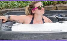 A woman with blonde hair and a tattoo on their shoulder leaning over the edge of a black hot tub with bubbles.