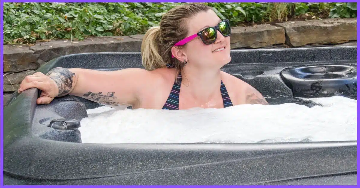 A woman with blonde hair and a tattoo on their shoulder leaning over the edge of a black hot tub with bubbles.