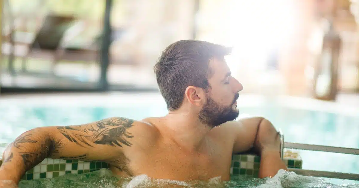 A man with a bird tattoo on his shoulder leaning on the edge of an indoor hot tub.