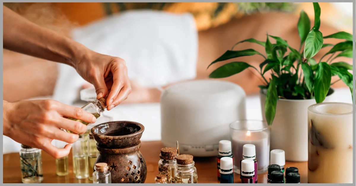 A photo of a person’s hand pouring essential oil into a diffuser on a wooden table with various bottles of essential oils, a white diffuser, a candle, and a plant.