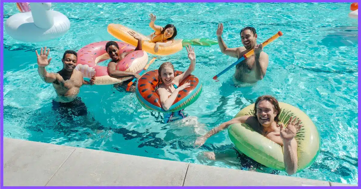 A group of people in a swimming pool with colorful fruit-shaped floaties, waving and smiling. (Particles in Your Pool)