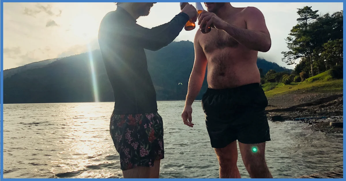 Two people standing on a rocky shore, clinking beer bottles with a mountain and lake in the background. (Wild Swimming)