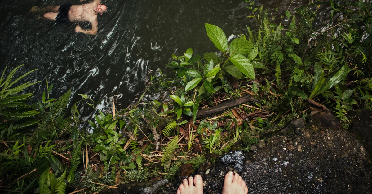 A person’s feet standing on a rock next to a body of water with plants surrounding it.