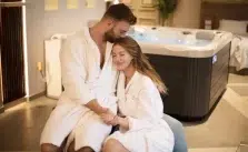 A couple in white bathrobes kissing passionately in front of a black hot tub. The room has a high ceiling and modern decor. The couple’s faces are blurred out for privacy reasons.