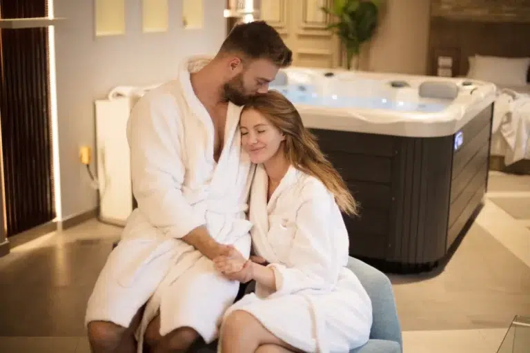 A couple in white bathrobes kissing passionately in front of a black hot tub. The room has a high ceiling and modern decor. The couple’s faces are blurred out for privacy reasons.