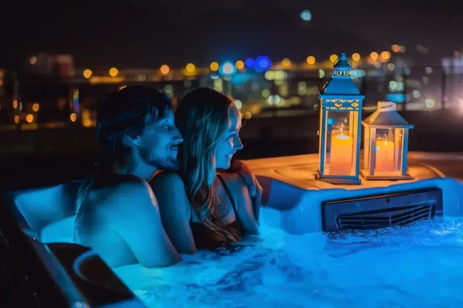 Two people in a hot tub with a lantern on the edge and a city skyline in the background. (Hot Tub Makeouts)