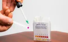 The person is testing the hot tub water alkalinity. A box of pH-Fix 7.9-9.8 indicator strips is visible in the background.