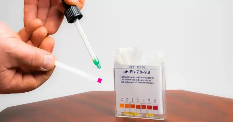 The person is testing the hot tub water alkalinity. A box of pH-Fix 7.9-9.8 indicator strips is visible in the background.