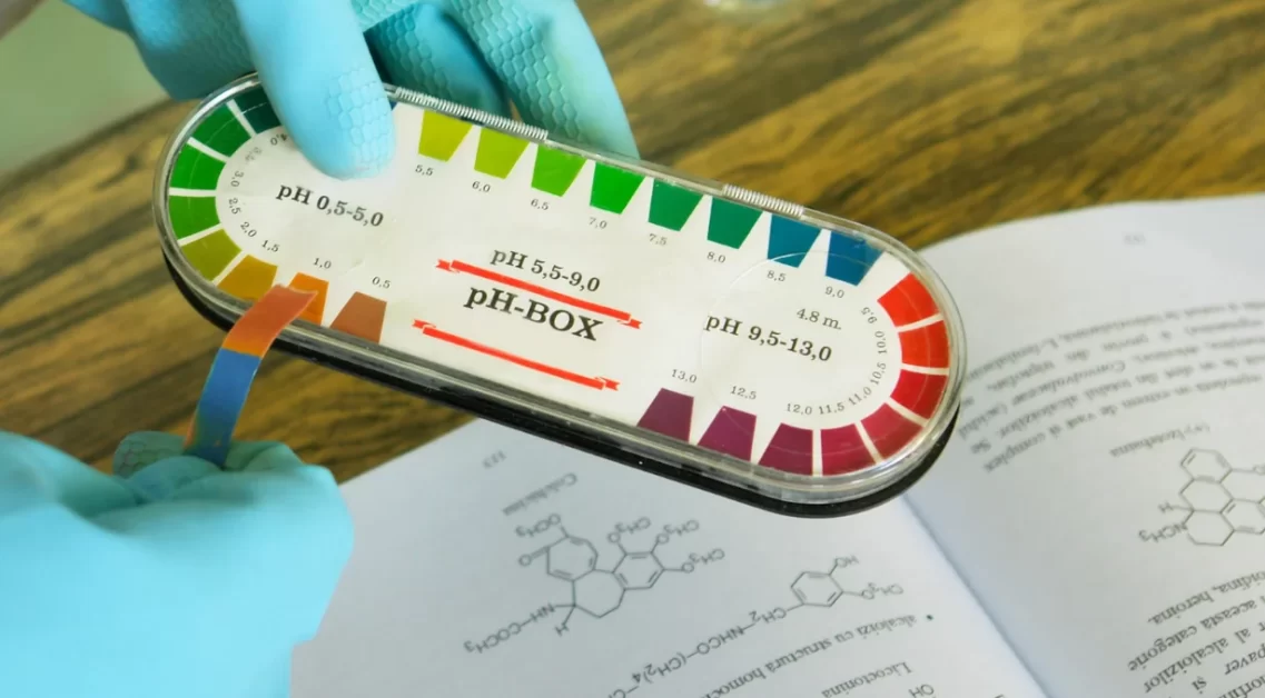 A person wearing blue gloves is holding a pH strip and a pH color chart over an open chemistry book.