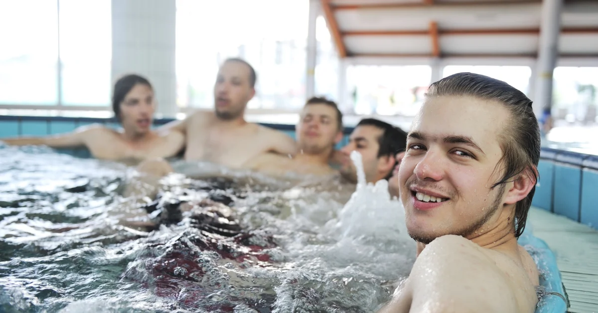 A group of young people enjoying a relaxing swim in an indoor pool. The image shows a smiling man in the foreground, with others visible in the background.