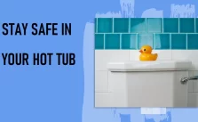 A white hot tub is set against a wall with blue tiles, accompanied by a yellow rubber duck perched on the edge. The text “STAY SAFE IN YOUR HOT TUB” is displayed prominently to the left.