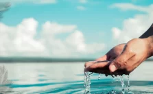 A close-up image of a person’s cupped hands scooping up clear water from a large body of water, with water droplets falling from the hands back into the body of water. The background shows a blurred blue sky with light clouds.