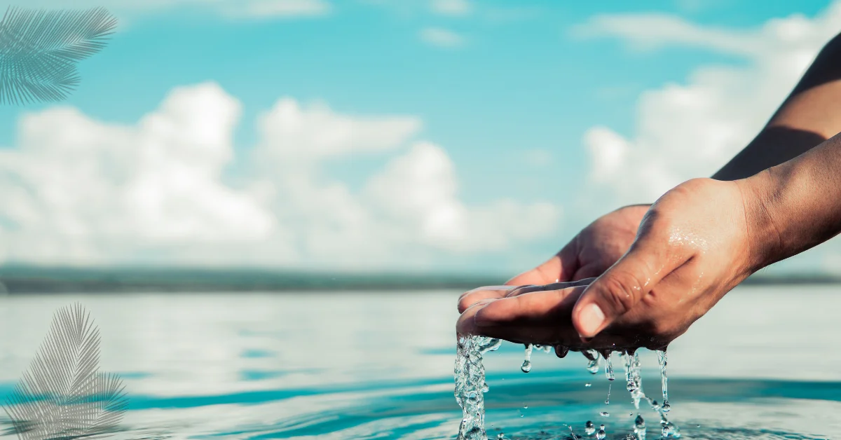 A close-up image of a person’s cupped hands scooping up clear water from a large body of water, with water droplets falling from the hands back into the body of water. The background shows a blurred blue sky with light clouds.