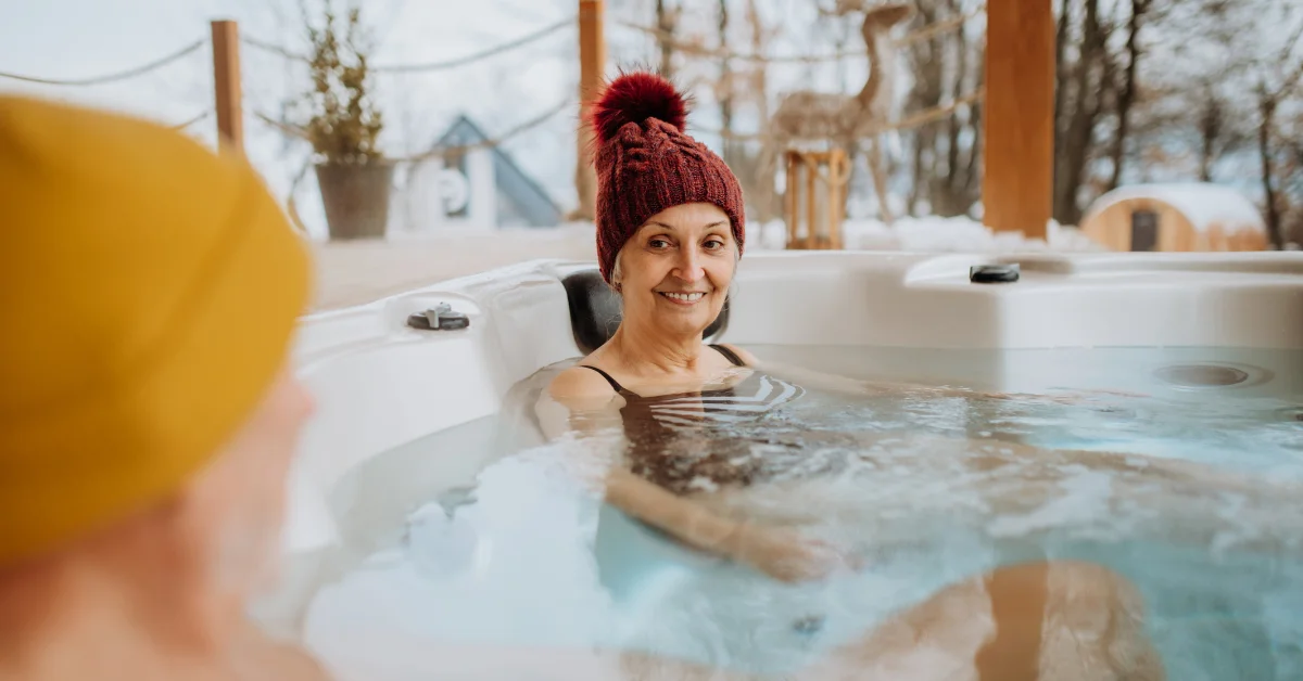 Woman with red knitted hat relaxing in a steaming hot tub outdoors in winter, another person with yellow cap seen from behind.