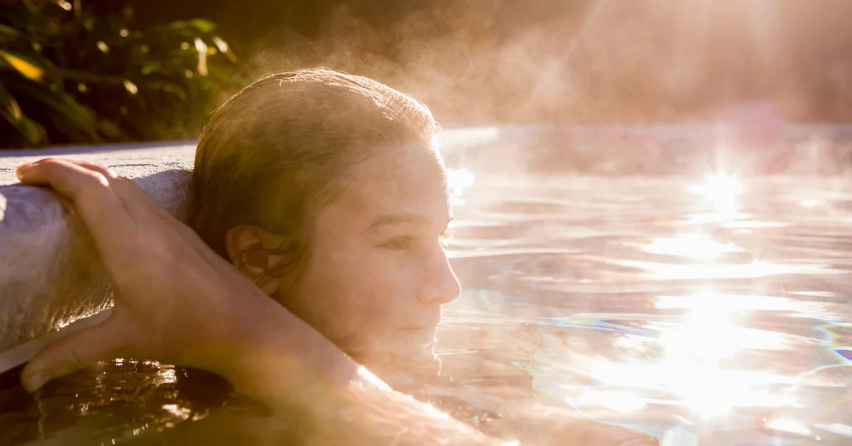 A woman is seen relaxing in a hot tub with their head resting on the edge, partially obscured by a brown rectangular censor bar over the face for privacy. The sun is shining brightly, creating a warm glow and reflecting off the water’s surface.