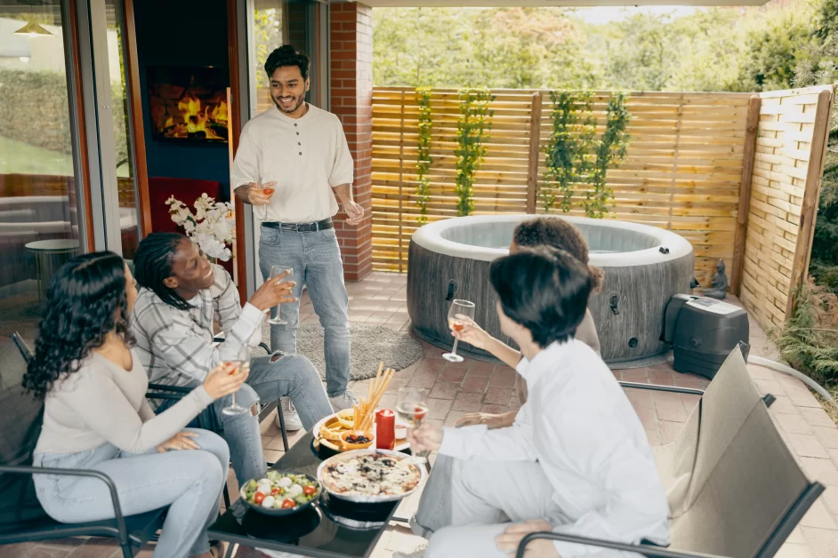 Four individuals are gathered on a patio with a inflatable hot tub in the background. One person stands holding a drink, while three others are seated around a table filled with various dishes. The setting appears to be a casual outdoor gathering.