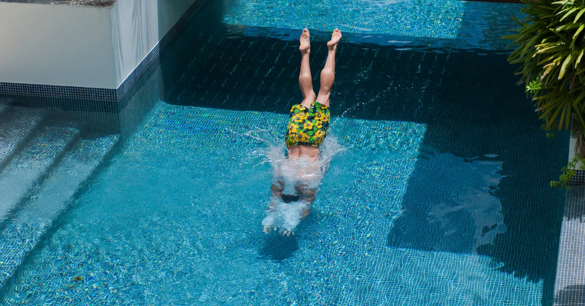 A person with their legs raised is partially submerged in a plunge pool with blue mosaic tiles, surrounded by a pool deck.