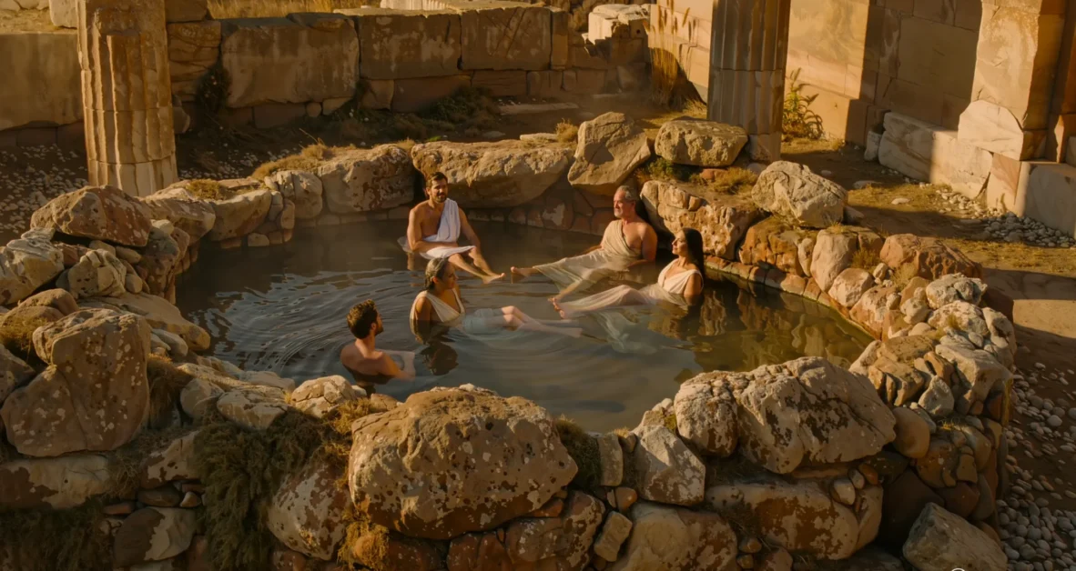 A historical hot water bathing scene set in ancient times, featuring a natural stone hot spring surrounded by rugged rocks and soft steam rising into the air.