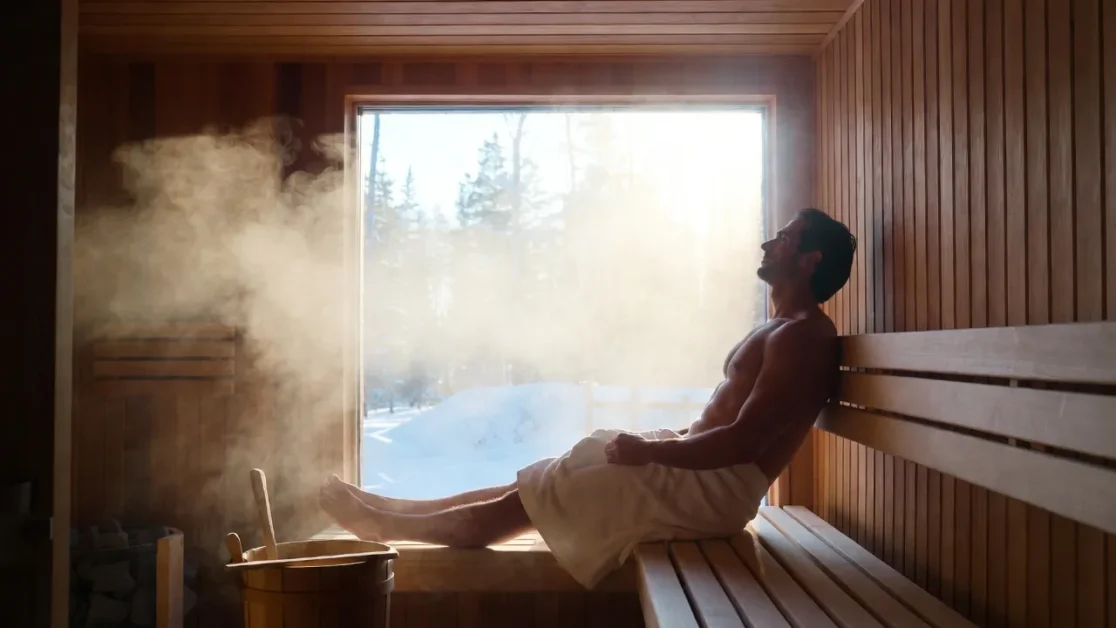 A warm wooden sauna interior with soft steam rising, a person sitting relaxed with a towel, winter scenery visible outside a small window.